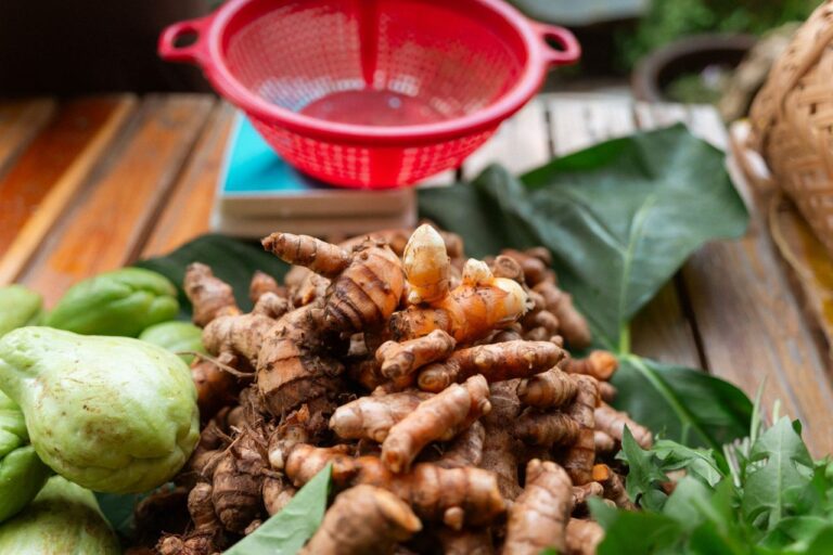 A vibrant display of fresh turmeric roots with leafy greens and chayote on a wooden table.