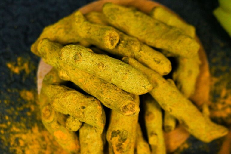 Изсушени корени от куркума в дървена купа, поръсени с жълт прах от куркума, върху тъмна повърхност.