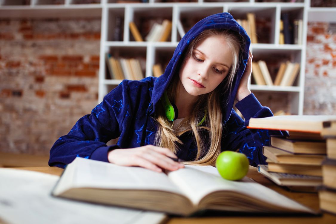 Free Teenage girl in blue hood reads in library, headphones and apple by her side. Stock Photo